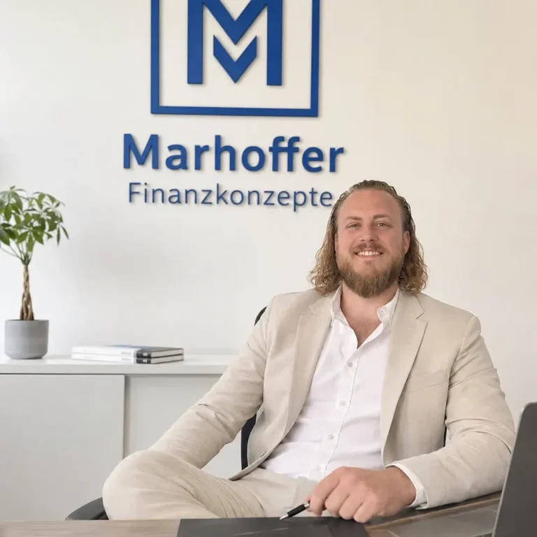 Inhaber Alexander Marhoffer sitzt lächelnd im Stuttgarter Büro vor dem Logo von Marhoffer Finanzkonzepte an der Wand. Alexander Marhoffer: deine erste Adresse für eine lückenlose Gesundheitsabsicherung